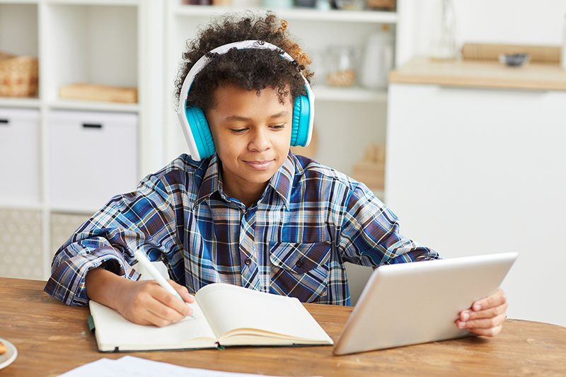 Young tween with headphones on, writing in a notebook and looking at tablet