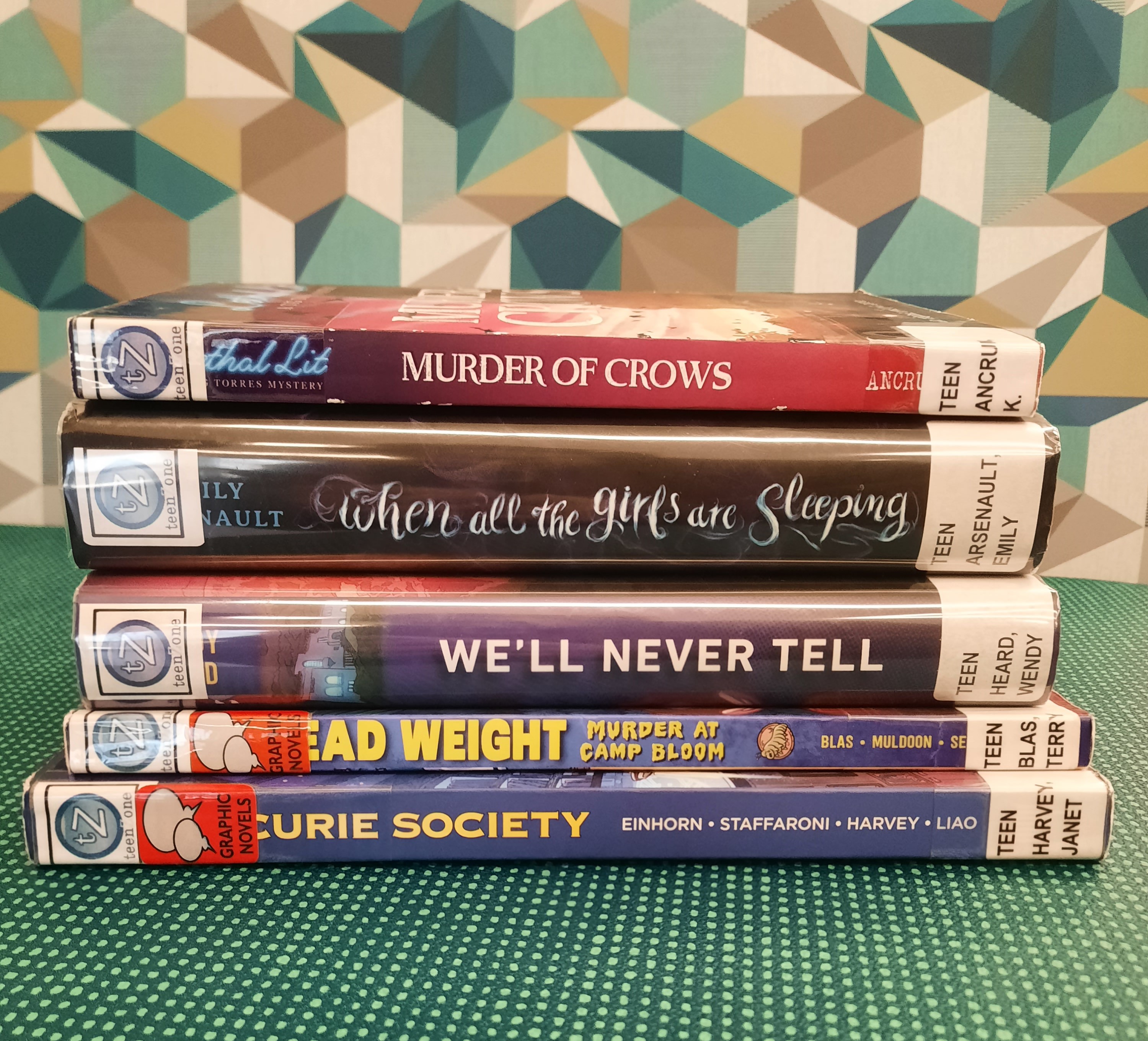 2025 December Book Stack Book Stack showing the following titles: "Murder of Crows," "When All the Girls are Sleeping," "We'll Never Tell," "Dead Weight: Murder at Camp Bloom," and "The Curie Society"